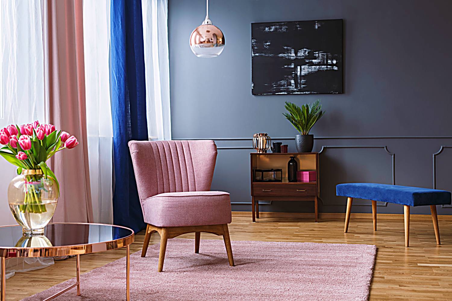 pink armchair standing on a rug and under a lamp in spacious living room interior, next to a table with flowers and in front of a shelf next to a grey wall with dark painting