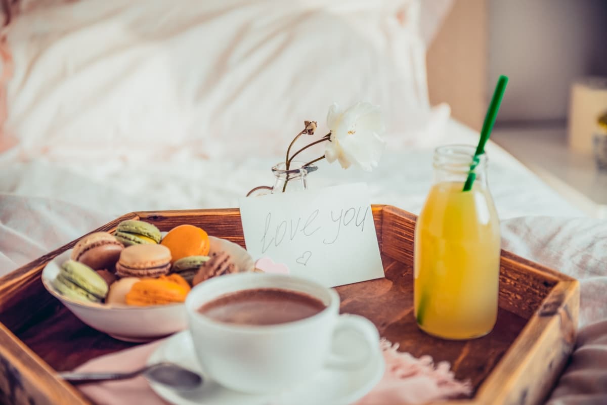 Breakfast in bed prepared for Valentine's Day, with a plate of macarons, a cup of coffee, a glass of orange juice, and a sweet note.